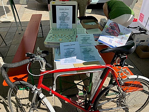 Rotes Fahrrad vor einem Tisch mit Ultraschallgerät, Infomaterialien und Urkunden.