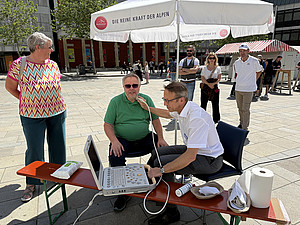 Arzt untersucht bei öffentlicher Aktion auf dem Neupfarrplatz die Halsschlagader eines Mannes per Ultraschall, mehrere Passanten schauen zu.